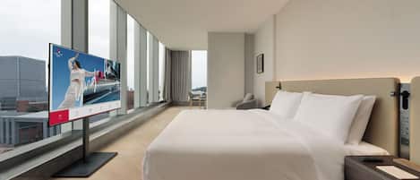 Habitación de hotel con una cama grande, un escritorio, una silla, un televisor y vistas a la ciudad a través de amplios ventanales.