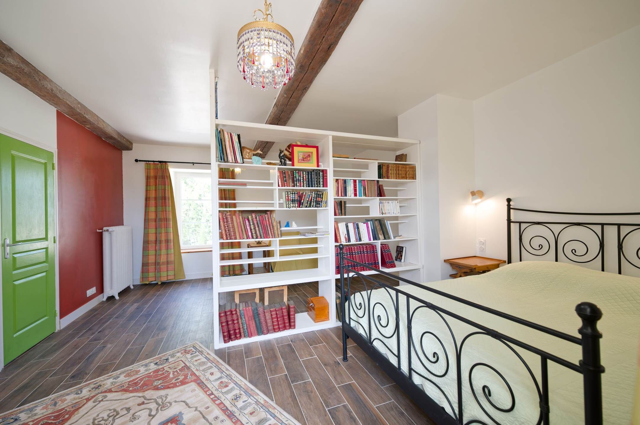 A bedroom with a bed, a white bookshelf, a radiator, a green door, and a window with curtains.
