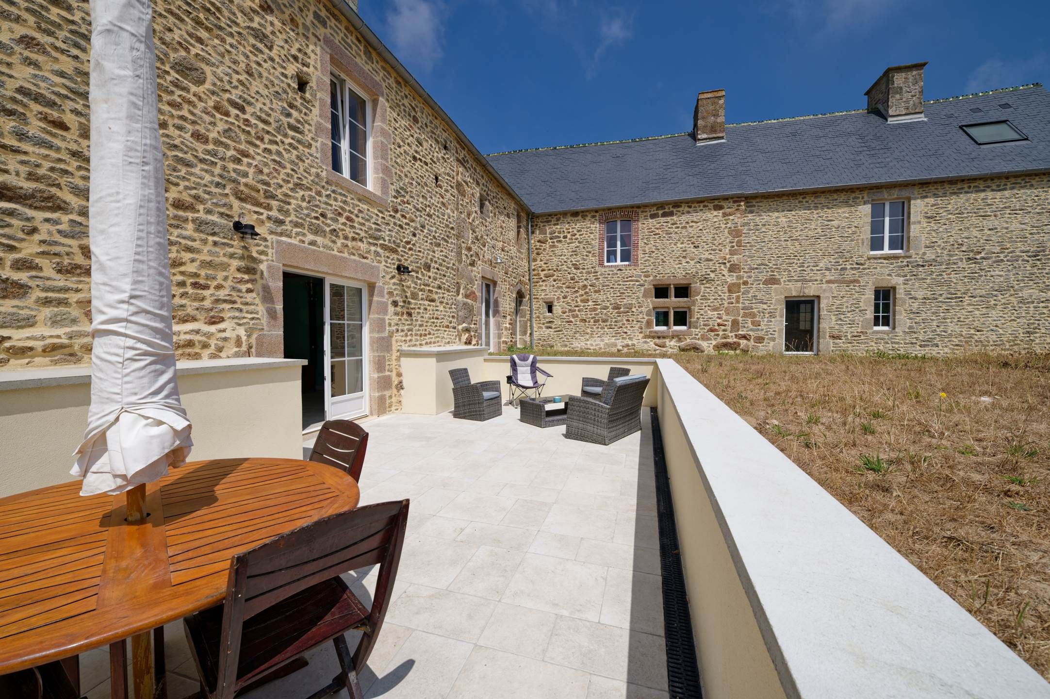 A stone building with a patio area featuring a wooden table and chairs, a white wall, and a clear sky.