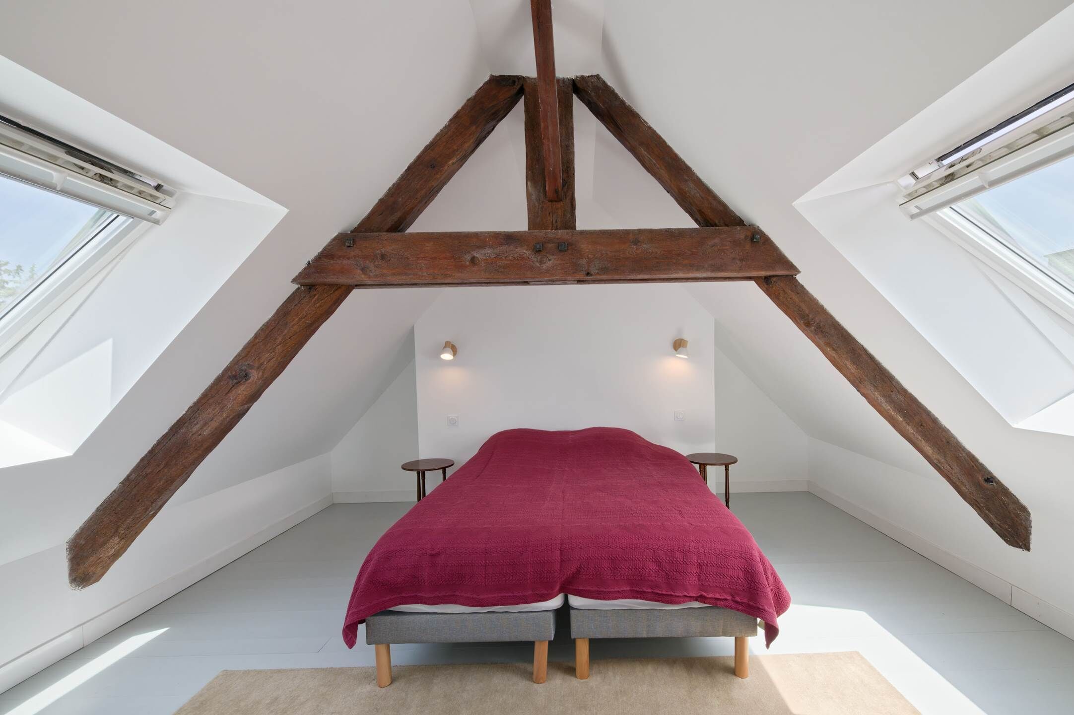 A bedroom with a sloped ceiling, wooden beams, a bed with a red blanket, and two wall-mounted lights.
