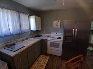 A compact kitchen with a refrigerator, stove, sink, and a window with curtains. - Affordable 3-Bedroom Stay in Wailuku, Maui (Wailuku)