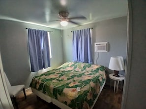 Quarto com cama, ventilador de teto, janela com cortinas, abajur e ar condicionado fixado na parede.