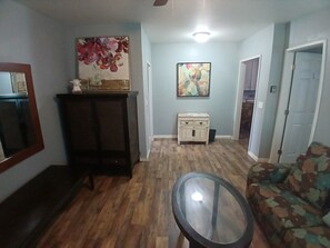 Una sala de estar con piso de madera, una mesa de centro de vidrio, un sofá, un mueble aparador y dos cuadros en la pared.