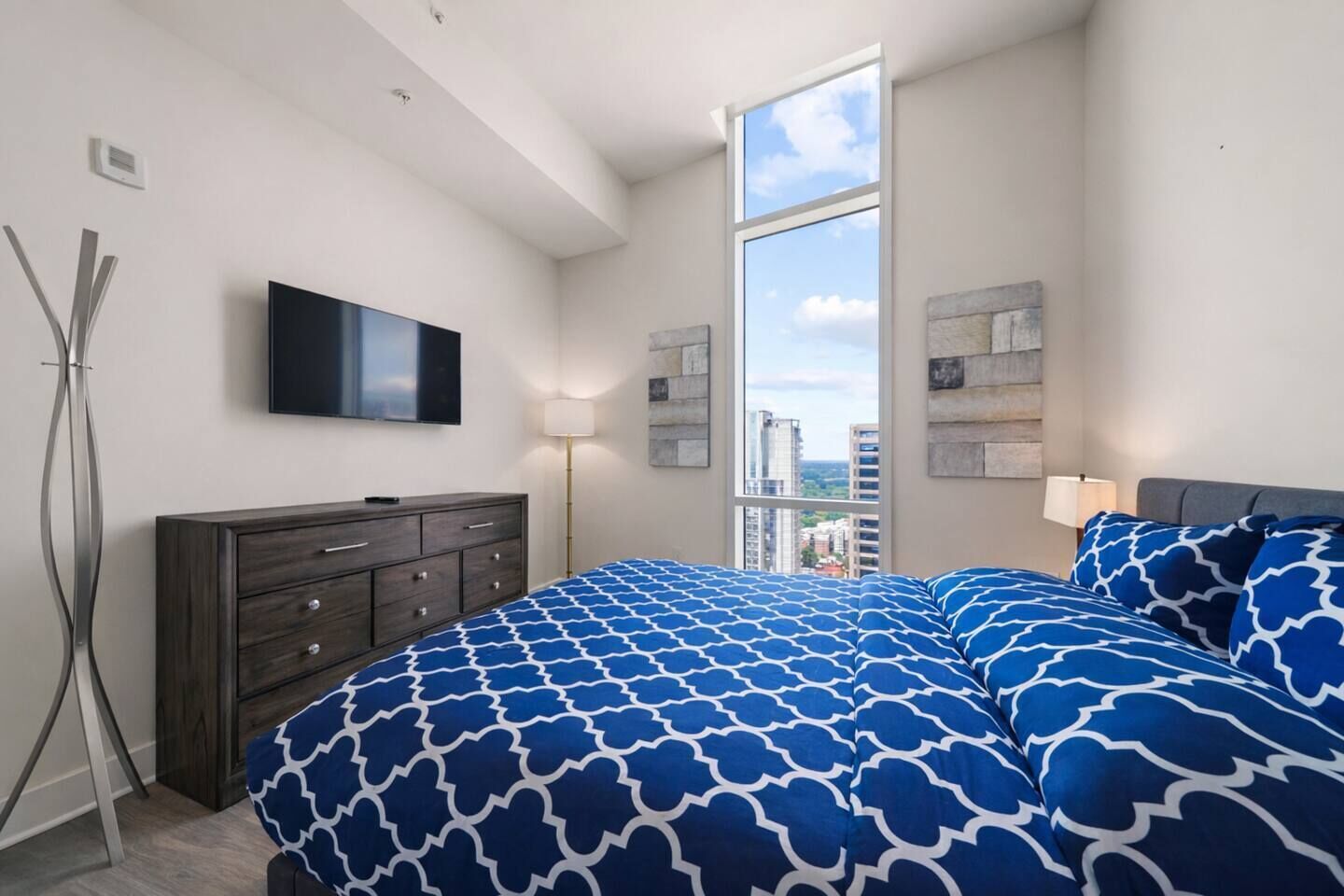 A bedroom with a large bed, a flat-screen TV, a dresser, and a window with a city view.