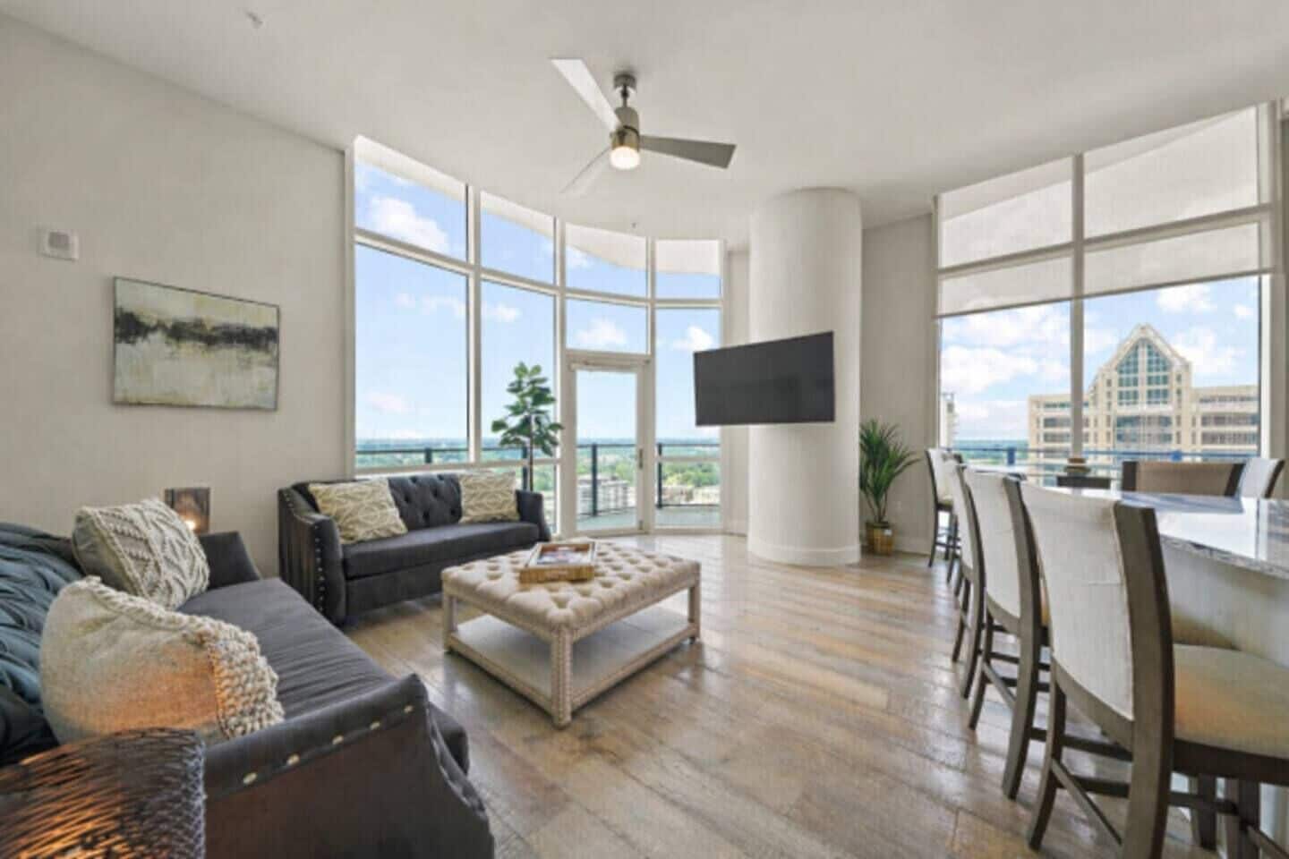 A spacious living room with a large window, a ceiling fan, a flat-screen TV mounted on a pillar, and a view of the ocean.