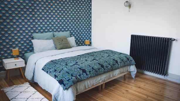 Um quarto com papel de parede estampado, uma cama com colcha florida, um abajur, um radiador e piso de madeira.