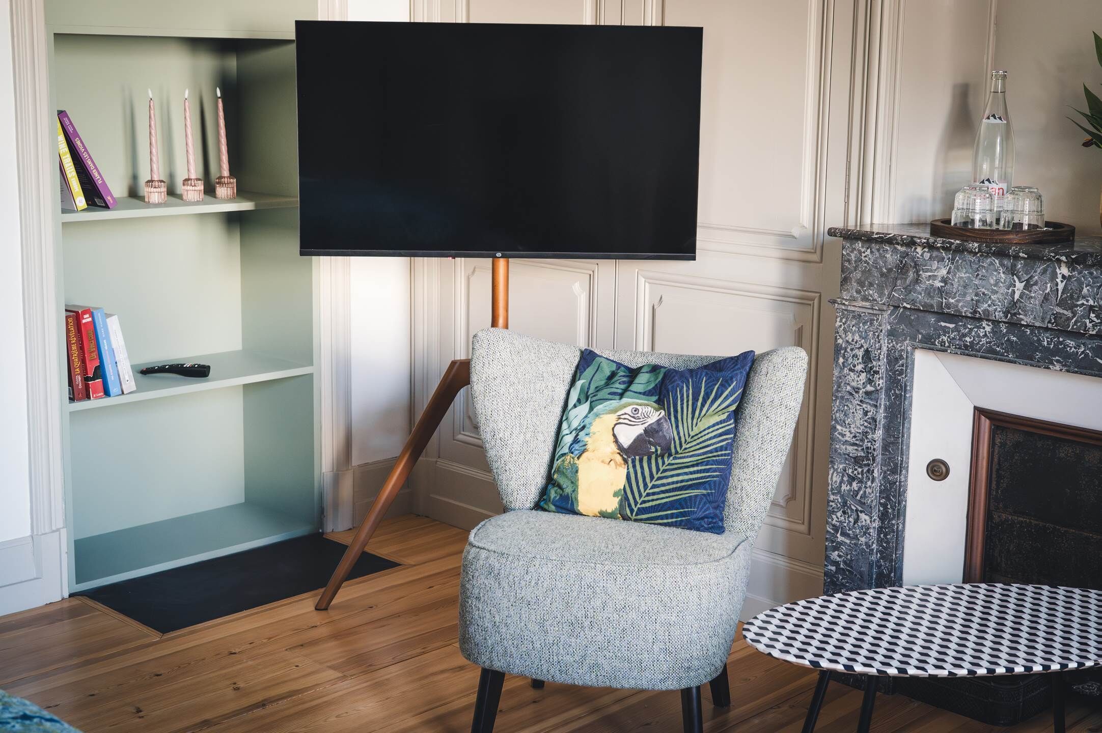 Ein Wohnzimmer mit Flachbildfernseher auf einem Standfuß, einem gemusterten Sessel mit Zierkissen, einem kleinen runden Tisch und einem Bücherregal mit Büchern und Kerzen.