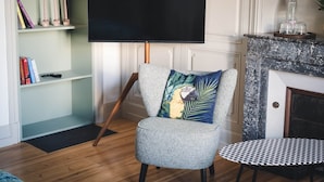 Uma sala de estar com televisão de tela plana em um suporte, uma poltrona estampada com uma almofada decorativa, uma pequena mesa redonda e uma estante com livros e velas.