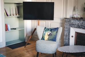 A living room with a flat-screen TV mounted on a stand, a patterned armchair with a decorative pillow, a small round table, and a bookshelf with books and candles.