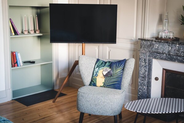 A living room with a flat-screen TV mounted on a stand, a patterned armchair with a decorative pillow, a small round table, and a bookshelf with books and candles.