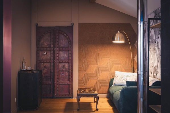 A room with a large wooden door, a herringbone-patterned wall, a floor lamp, a bed with a floral pillow, and a small table.