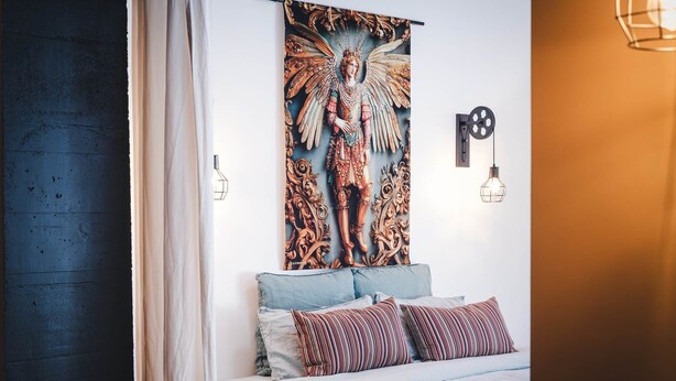 Een slaapkamer met een bed, kussens, een wandtapijt en een wandlamp.
