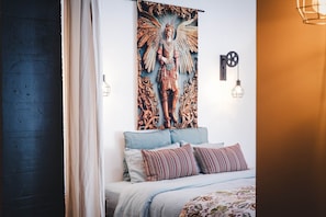 A bedroom with a bed, pillows, a wall tapestry, and a wall-mounted light fixture.