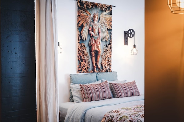 Un dormitorio con cama, almohadas, un tapiz en la pared y una lámpara fijada a la pared.
