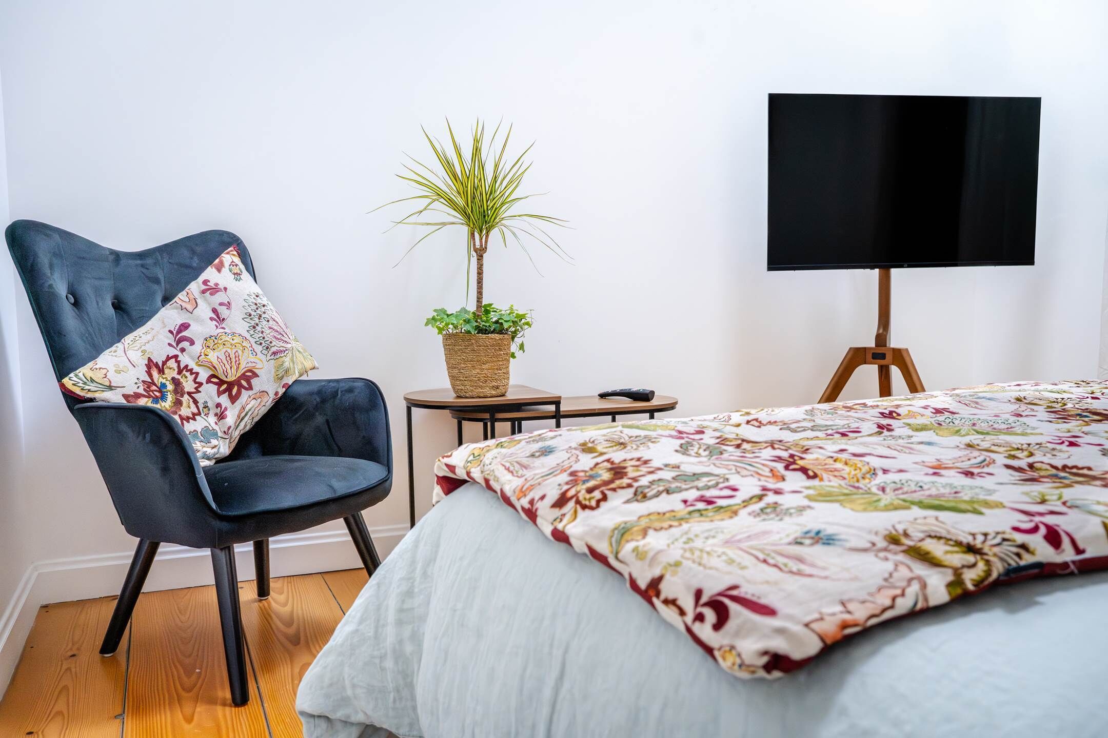 A bedroom with a bed, a chair, a small table, a plant, and a TV on a stand.