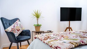 Een slaapkamer met een bed, een stoel, een klein tafeltje, een plant en een televisie op een standaard.