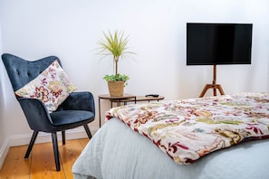 Un dormitorio con cama, una silla, una mesita, una planta y un televisor sobre un mueble.