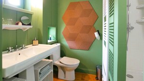 Een badkamer met een wit wastafelmeubel, toilet en douche. De wand is groen en er is een decoratieve houten wandversiering in een honingraatstructuur.