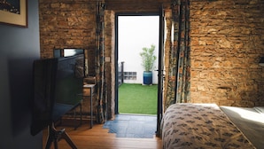 Un dormitorio con pared de piedra, una cama con ropa de cama floral, un escritorio con una silla y vistas a un césped verde a través de una puerta abierta.