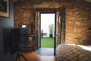 Une chambre avec un mur en pierre, un lit recouvert d’une literie à motifs floraux, un bureau avec une chaise, et une vue sur une pelouse verdoyante par une porte ouverte.