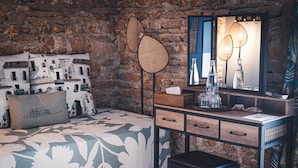 Un dormitorio con pared de piedra, una cama con ropa de cama estampada, una mesita de noche con espejo y una lámpara de pie.