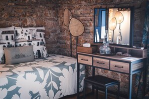 Une chambre avec un mur en pierre, un lit avec une literie à motifs, une table de chevet avec un miroir et une lampe de sol.