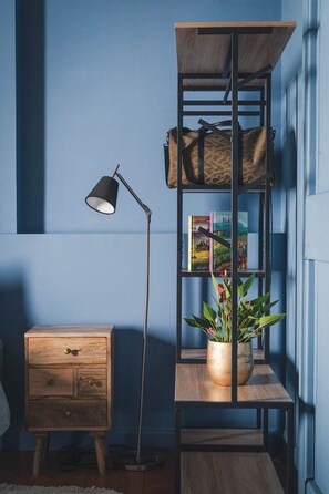 A room with a wooden nightstand, a floor lamp, a bookshelf with a plant, and a blue wall. - Les Jardins de Cormatin Room (FUISSE)