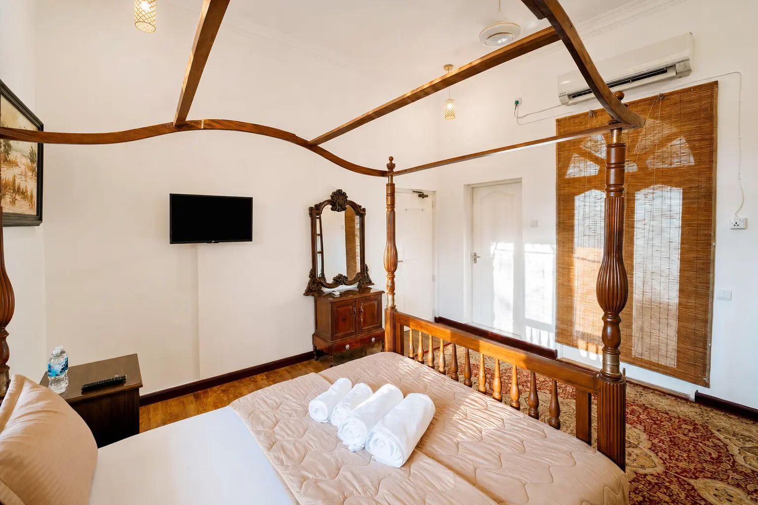 A four-poster bed with rolled towels, a wooden dresser, a flat-screen TV, and a large window with bamboo blinds.