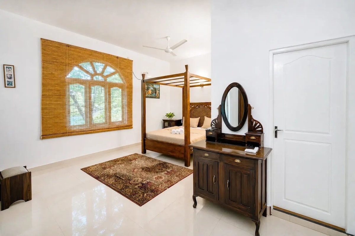 A bedroom with a four-poster bed, a large mirror, a wardrobe, and a window with bamboo blinds.