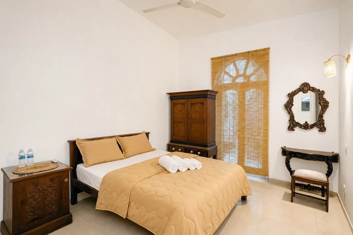 A hotel room with a bed, a wooden nightstand, a wardrobe, a mirror, and a window with blinds.