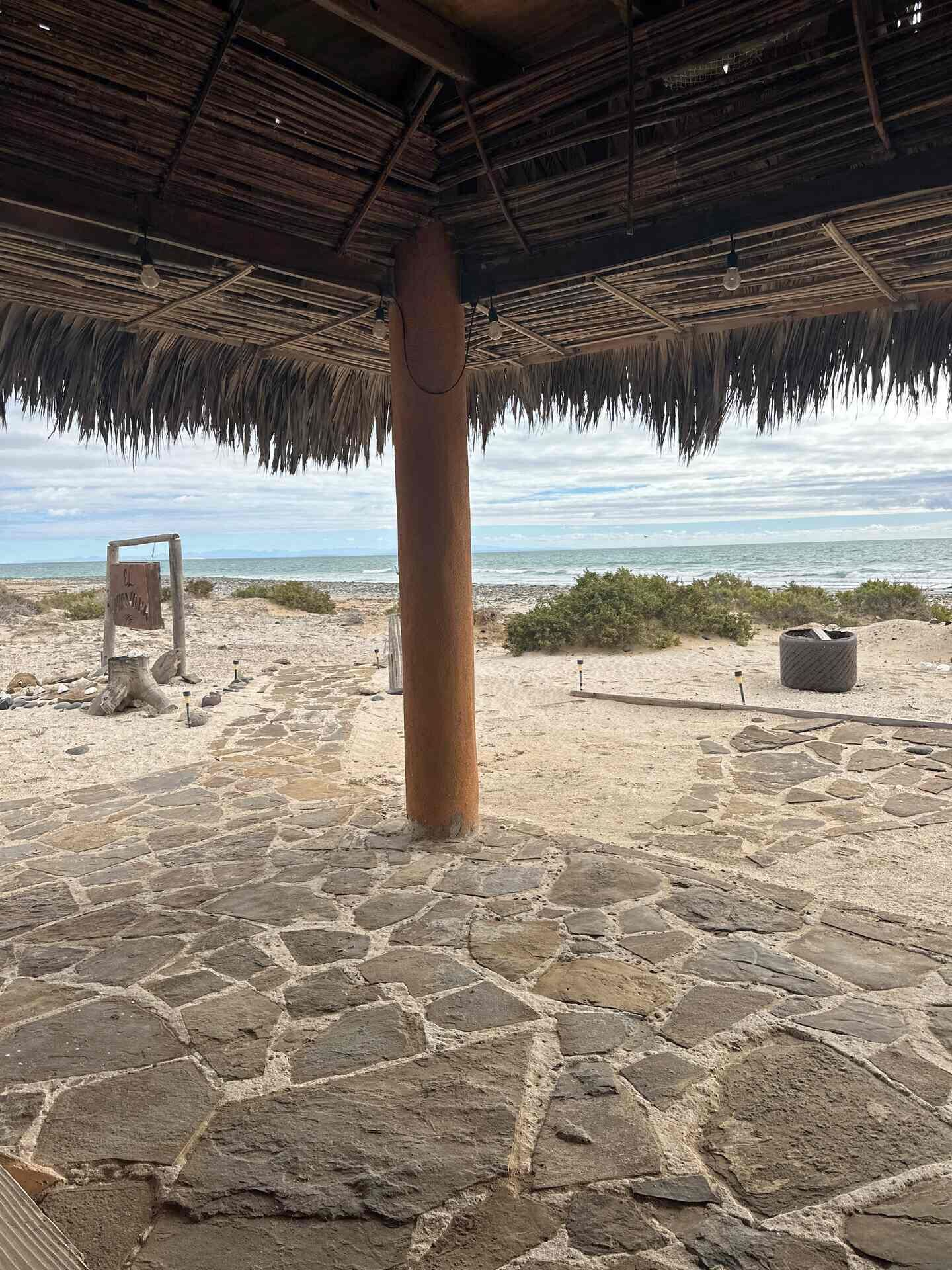 Een strand met een verhard terras, een rieten gebouw en een bord.