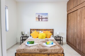 Une chambre à coucher avec un lit, des oreillers, une table de chevet, une lampe, un tableau au mur et une fenêtre avec des stores.