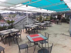 Un patio exterior con mesas y sillas, una zona cubierta y vista a la ciudad.