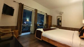 A hotel room with a bed, a desk, a chair, a television, and a view of the city at night. - Eden Paradisee (Ootacamund)
