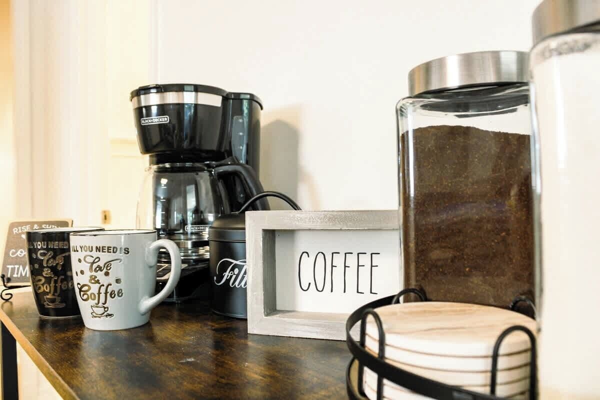 A coffee-making setup with a coffee maker, mugs, a coffee-themed sign, and a candle holder.