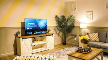 Ein Wohnzimmer mit Flachbildfernseher, einer Couch, einem Couchtisch, einem gemusterten Hocker und einer Zimmerpflanze sowie einer Lampe.