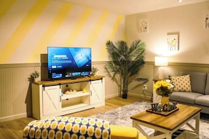 A living room with a flat-screen TV, a sofa, a coffee table, a patterned ottoman, a potted plant, and a lamp. - Quiet Home w/ Sunroom & Fire Pit Near Downtown (Jeffersonville)