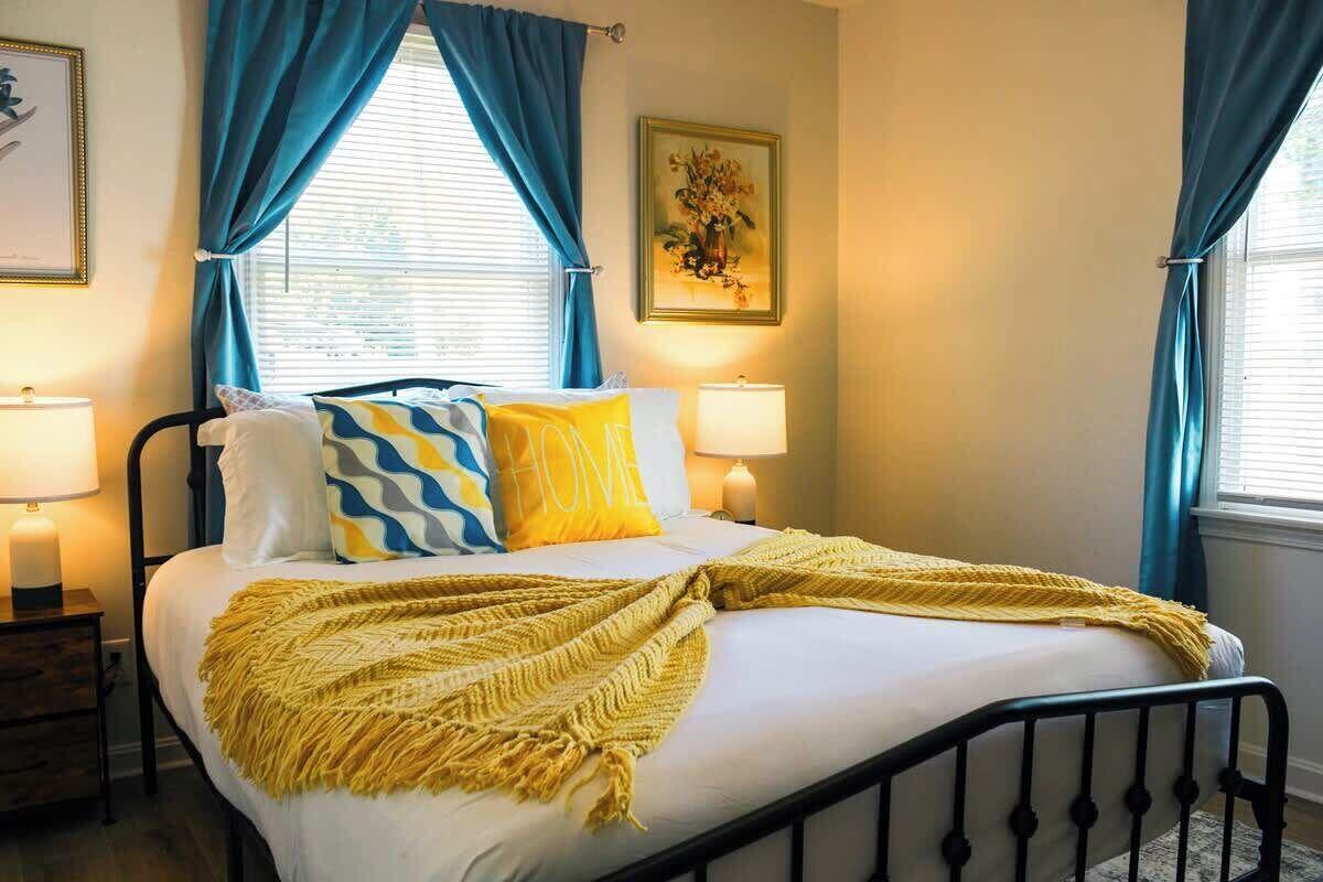 A bed with a yellow throw blanket, two bedside lamps, and a painting on the wall.
