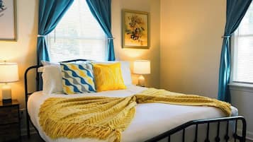 A bed with a yellow throw blanket, two bedside lamps, and a painting on the wall.