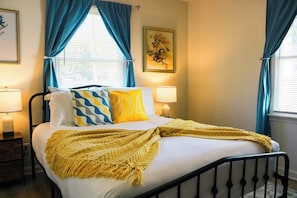 A bed with a yellow throw blanket, two bedside lamps, and a painting on the wall. - Quiet Home w/ Sunroom & Fire Pit Near Downtown (Jeffersonville)