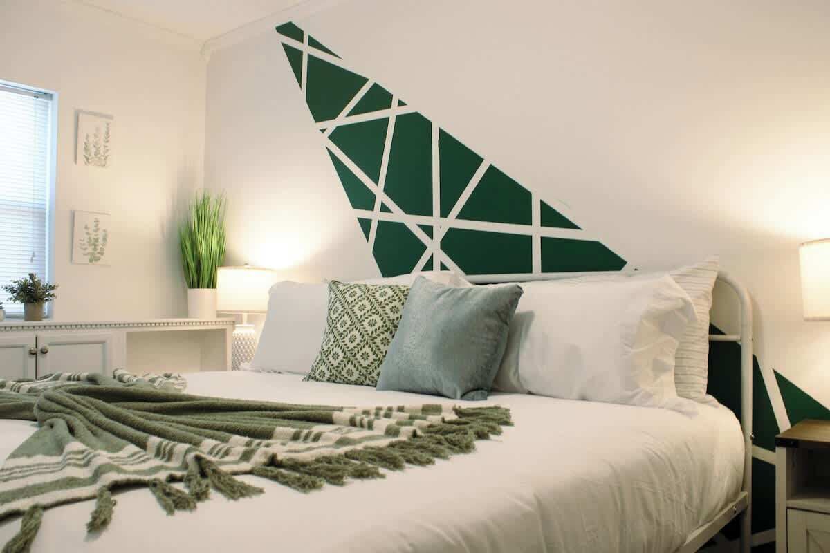 A bedroom with a bed, pillows, a green wall decoration, a bedside table, a lamp, and a window.