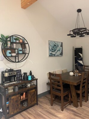 A dining area with a wooden table, chairs, a coffee station, a wall-mounted shelf with kitchenware, and a painting of a rose. - luxury ranch home in the mountains in Quad country near Manning Park ski resort. (Hope)