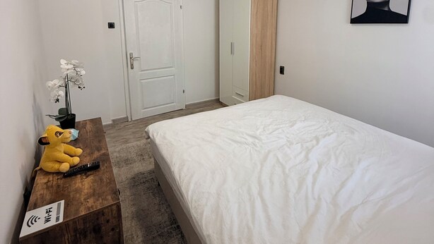 Un dormitorio con una cama, un aparador de madera, un televisor montado en la pared, una puerta y un cuadro enmarcado.