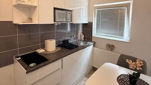 Cocina moderna con armarios blancos, microondas y lavamanos. Hay una mesa de comedor con sillas y una ventana con persianas.
