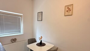 Un pequeño comedor con una mesa blanca, dos sillas grises, un jarrón con flores, un cuadro enmarcado en la pared y una ventana con persianas.