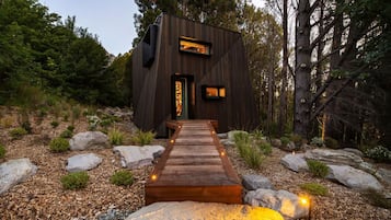 A modern wooden cabin with a wooden deck leading to the entrance, surrounded by rocks and trees.