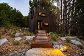 A modern wooden cabin with a wooden deck leading to the entrance, surrounded by rocks and trees.