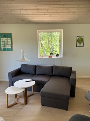 Ein modernes Wohnzimmer mit einem Ecksofa, einem kleinen weißen Tisch und einem Fenster mit Blick auf Bäume.
