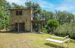 Ein Steingebäude mit Holztür und Fensterläden, eine Pergola mit Tisch und Stühlen sowie eine Rasenfläche mit einer Bank.