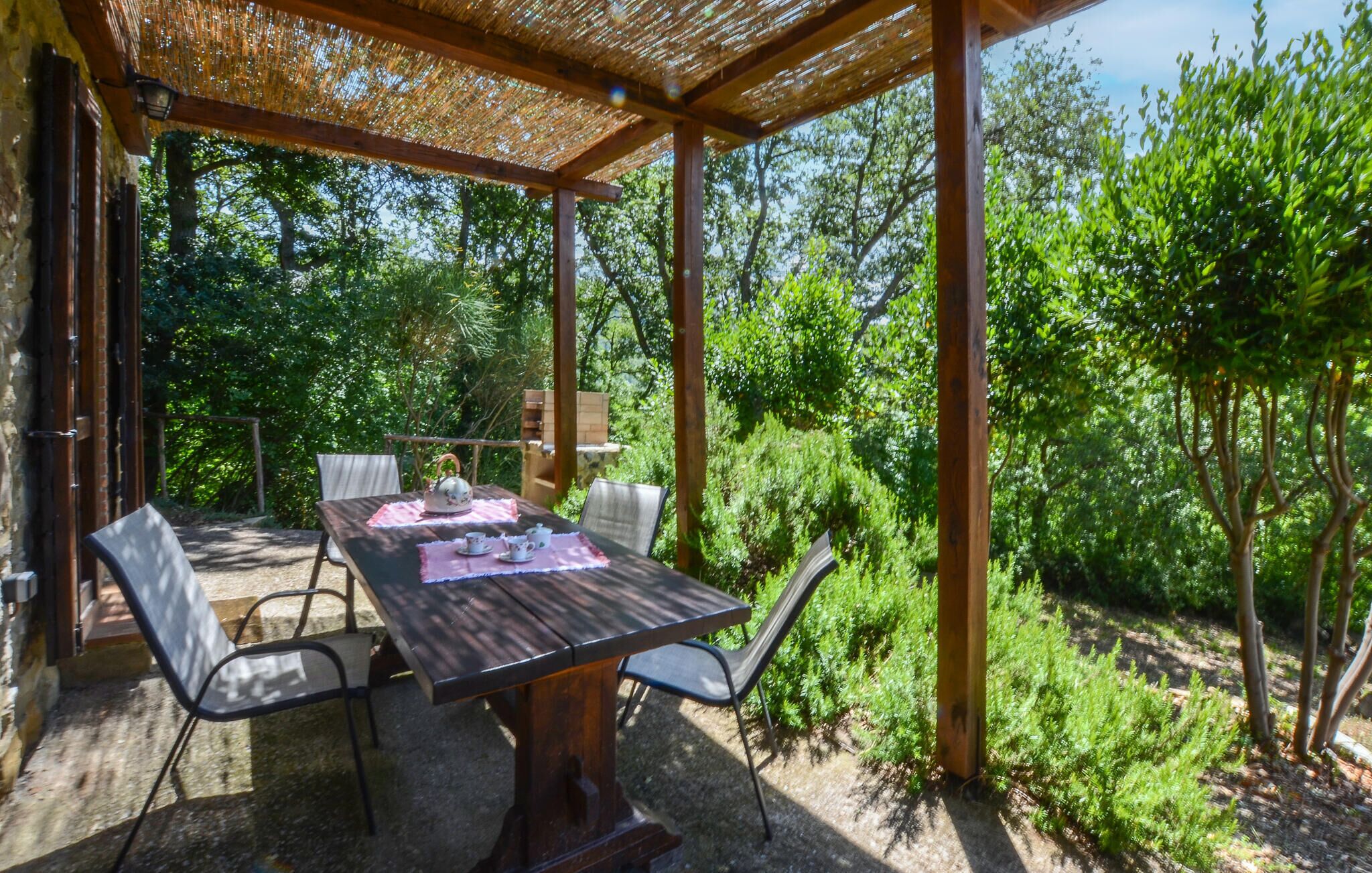 An outdoor patio with a wooden table and chairs, surrounded by lush greenery and trees.